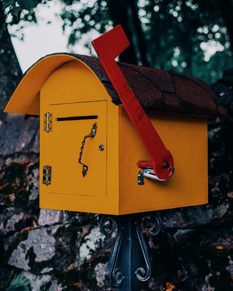 Bright yellow mailbox with red flag in a rustic outdoor setting, surrounded by greenery.